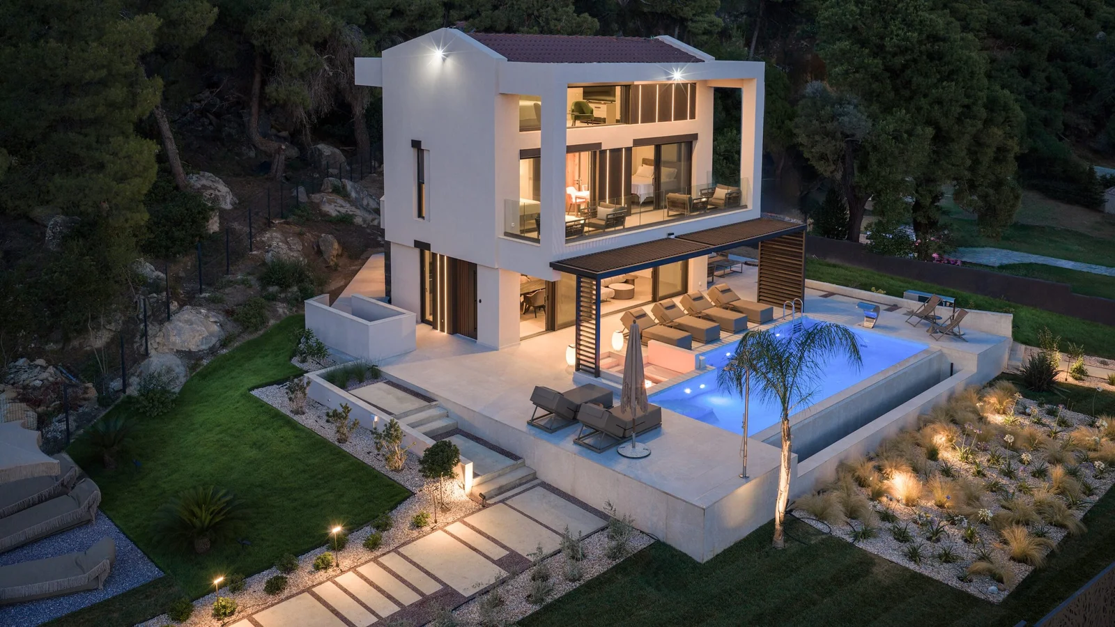 Villa Elyon lit at twilight, framed by pine forest with the sea beyond